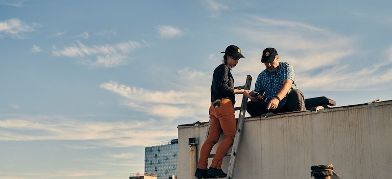 A woman and a man standing on a rooftop, gazing intently at an unseen object